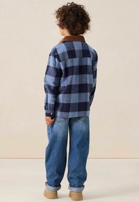 Blauw en marineblauw geruit overhemd met een bruine corduroy kraag, gecombineerd met losse blauwe jeans en beige schoenen. Effen achtergrond.