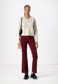 Lys grå cardigan med knapper, kombinert med burgunder kordfløyelsbukser med vide ben og svarte kraftige støvler. Modellen står mot en hvit bakgrunn.