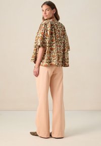 Bluse mit Blumenmuster in Erdtönen und weit geschnittenen, luftigen Ärmeln, kombiniert mit hellen Pfirsichfarbigen weiten Hosen und gemusterten Ballerinas. Neutraler Hintergrund.