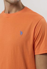 Oranžové bavlnené tričko s okrúhlym výstrihom a krátkymi rukávmi, na ľavej strane na prsiach s modrým vyšívaným logom.