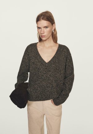 Femme portant un pull V en tissu texturé sombre, un pantalon beige, tenant une veste marron foncé pliée d'une main, l'autre main dans sa poche.