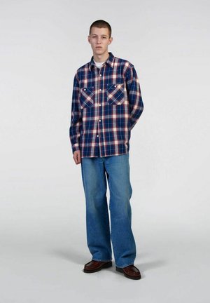 Camicia di flanella a scacchi in navy, rosso e bianco, con due tasche sul petto e chiusura a bottoni. Abbinata a jeans a gamba larga di un blu chiaro e scarpe marroni.