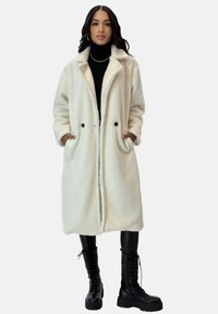 Witte, gestructureerde lange jas met voorste knoopsluiting, zijzakken en brede kraag. Draagt over een zwarte coltrui en legging, gecombineerd met zwarte laarzen.