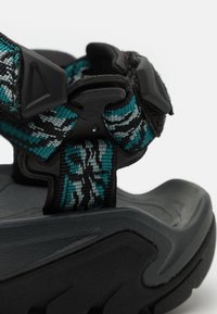Sandale noire avec une semelle texturée, dotée d'une sangle tressée aux motifs teal, gris et noir, sécurisée par une boucle en plastique.
