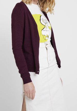 Femme portant un cardigan marron sur un t-shirt blanc avec un motif abstrait de visage, associé à une jupe blanche taille haute avec une fente à l'avant.
