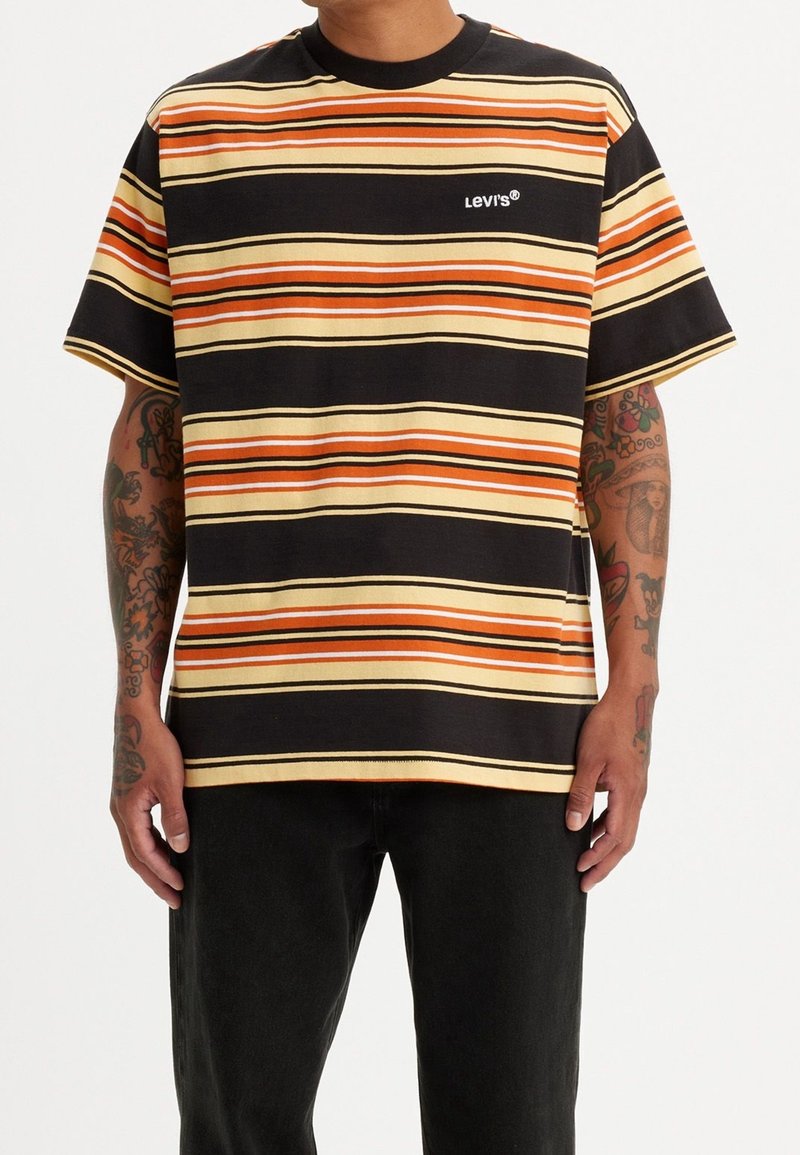 Homme portant un t-shirt Levi’s à manches courtes rayé noir, orange et crème avec des bras tatoués et un pantalon noir sur un fond clair.