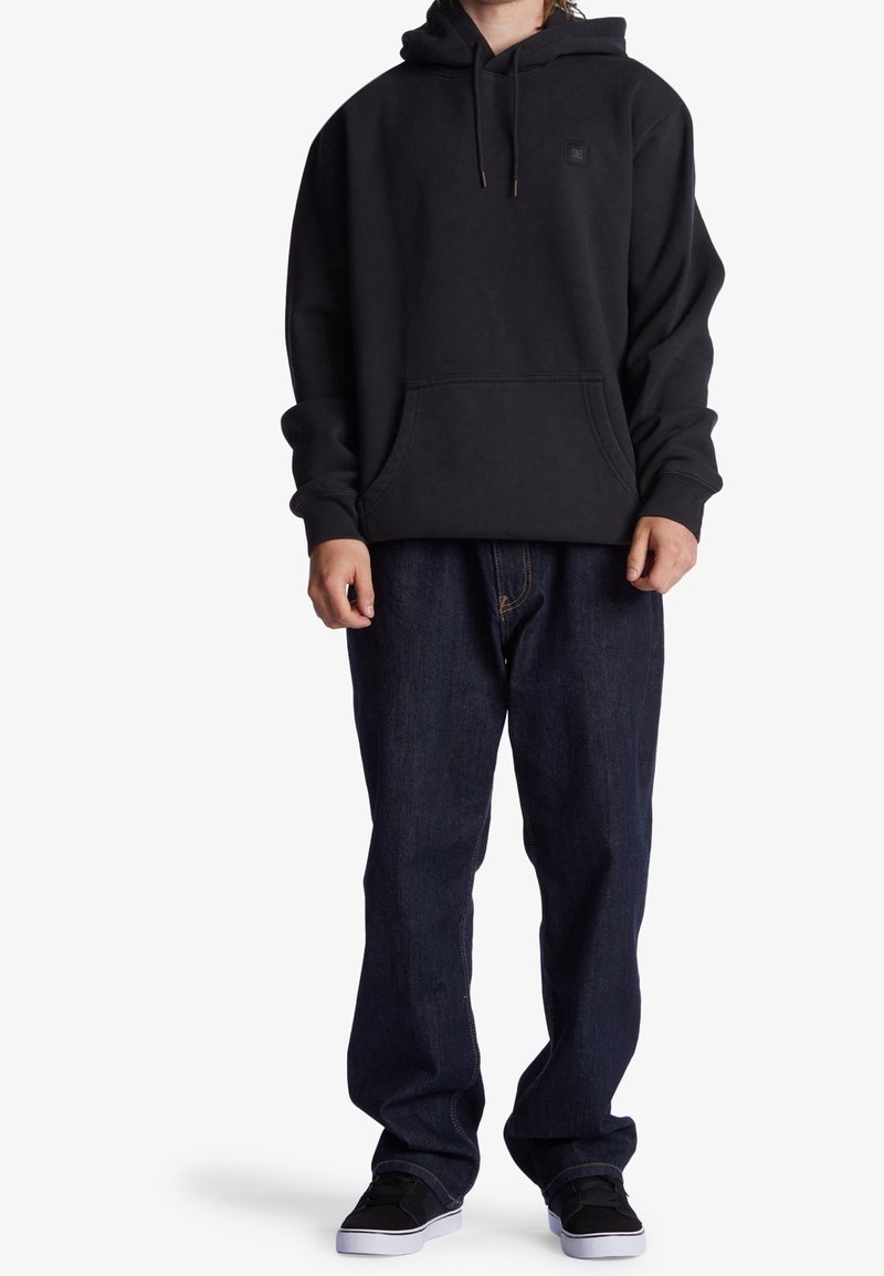 Un sweat à capuche noir avec une poche frontale et un cordon de serrage, associé à un jeans en denim noir ample et des baskets noires.