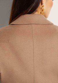 Bruine wolblend jas met een gestructureerde kraag, met een soepele textuur en een naaddetail in het midden aan de achterkant. Gouden oorbellen zichtbaar.