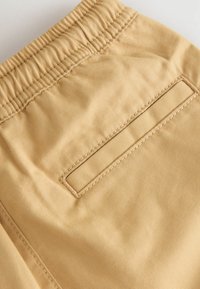 Lichtbeige shorts gemaakt van zachte stof met een elastische tailleband en een achterzak met gestikte details. Gladde textuur.