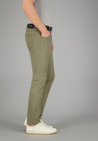 Olivgrüne Hose im Slim Fit mit Fronttaschen und gesticktem Akzent. Kombiniert mit weißen Sneakern und einem grauen T-Shirt.