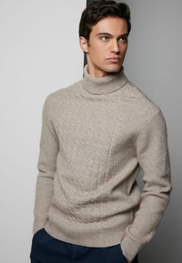 Beige stickad tröja med hög krage, med en texturerad diamantmönster framtill och ribbade ärmar och fåll.