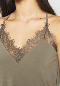 Camiseta sin mangas verde oliva con escote en V y intrincado encaje marrón en los bordes, confeccionada con una tela suave y tirantes finos.