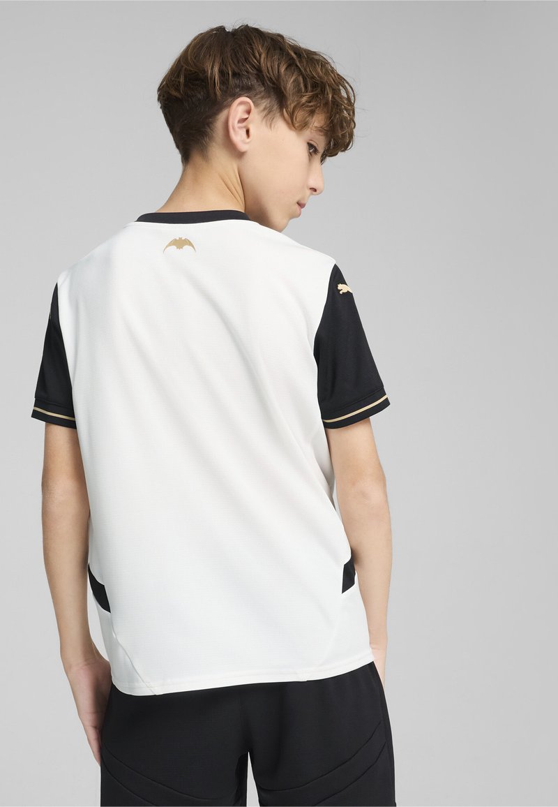 Camisa deportiva blanca y negra con cuello redondo, mangas cortas, logo de bate dorado en la parte trasera y acentos laterales negros. Textura suave.