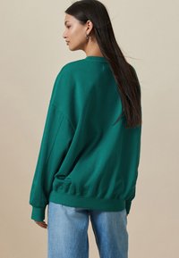 Zelená nadmerná mikina s rebrovanými manžetami, okrúhlym výstrihom a nazberaným spodným lemom. Kombinovaná s svetlomodrými džínsami. Jemná textúra, bavlnená zmes.