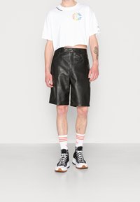 Svarta läderbermuda-shorter, vit kortärmad t-shirt med flerfärgad grafik, svarta högskurna sneakers och randiga strumpor.