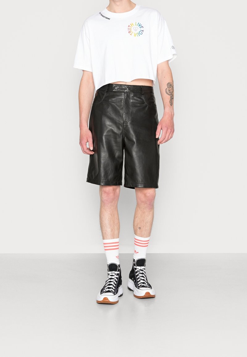 Svarta läderbermuda-shorter, vit kortärmad t-shirt med flerfärgad grafik, svarta högskurna sneakers och randiga strumpor.