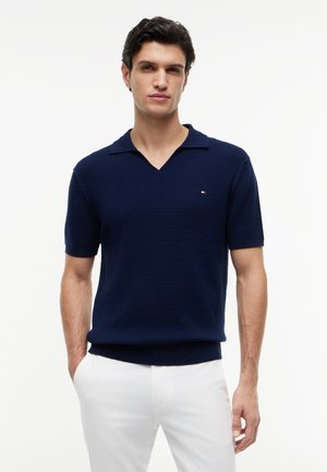 Mand iført en marineblå kortærmet strikket polo og hvide bukser, stående med den ene hånd i lommen.