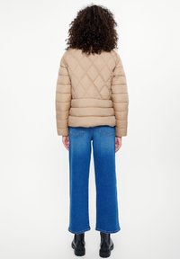 Beżowa pikowana kurtka puffer w diamentowy wzór, noszona na szerokie niebieskie dżinsy; czarne botki za kostkę uzupełniają strój.