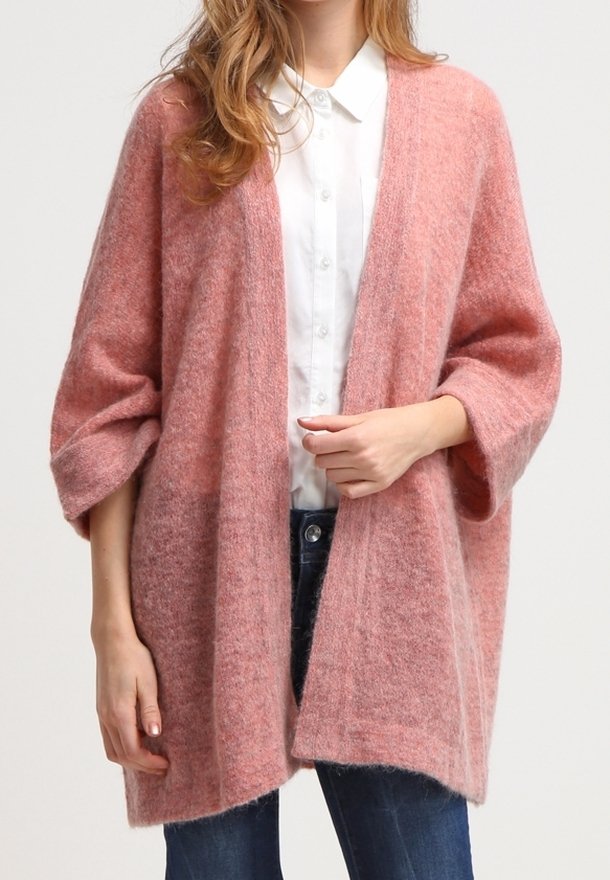Femme portant un cardigan en peluche rose doux par-dessus une chemise blanche à boutons et un jean bleu foncé, avec des manches amples et un design à l'avant ouvert.
