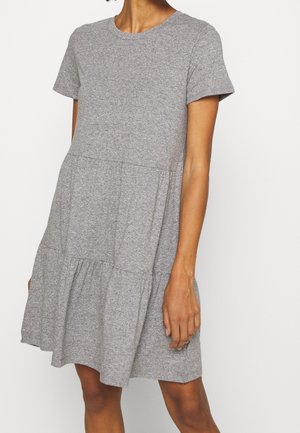 Femme portant une robe gris chiné à manches courtes et longueur genou avec une jupe à étages, debout sur un fond blanc.