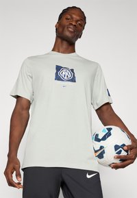 Ανοιχτό γκρίζο κοντομάνικο μπλουζάκι με μπλε και μαύρο γραφικό που αναγράφει "Interazionale Milano." Το μοντέλο κρατά μια μοτίβο ποδοσφαιρική μπάλα.