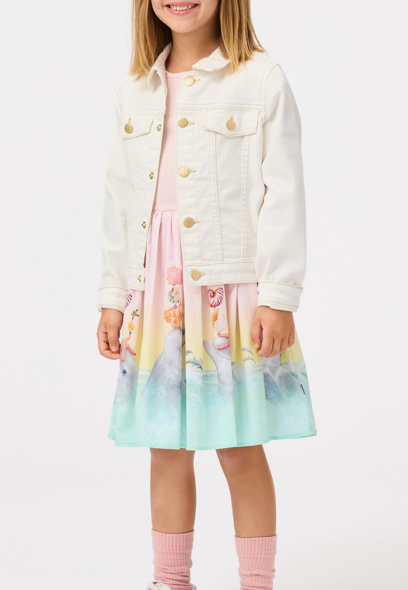 Weiße Jeansjacke mit goldenen Knöpfen, die über einem rosa Kleid getragen wird. Das Kleid zeigt einen bunten Stranddruck mit Delfinen und Meereslebewesen.