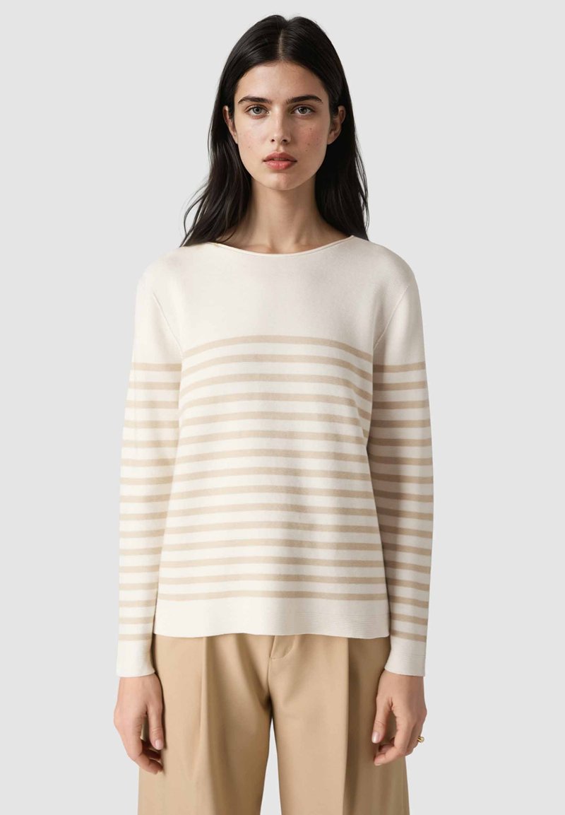Frau mit langen dunklen Haaren, die einen cremefarbenen und beigen langärmeligen Pullover mit Streifen und beige Hosen trägt, vor einem schlichten hellen Hintergrund.