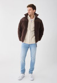 Brun fleecejakke med høy krage og kontrasterende brune skulderpaneler, båret over en lys beige hettejakke med glidelås, kombinert med lyseblå jeans og hvite joggesko.