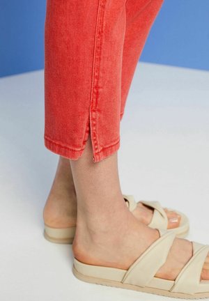 Jeans corta color corallo con spacchi laterali sull'orlo, abbinati a sandali beige con cinturini incrociati. Trama denim visibile sui pantaloni.