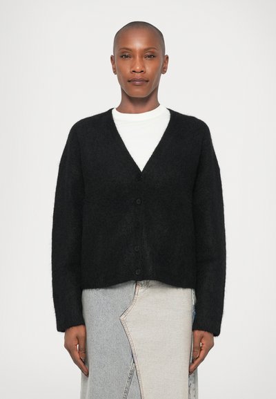 Cardigan nero peloso con scollo a V, maniche lunghe e cinque bottoni. Indossato sopra una camicia bianca e una gonna di jeans chiara.