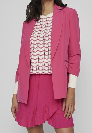 Kvinde iført en lyserød blazer og matchende flæset nederdel over en hvid striktrøje med pink bølgemønstre.