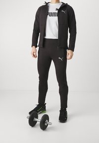 Conjunto atlético negro que incluye una chaqueta con cremallera y pantalones ajustados, hechos de un material suave, combinado con zapatillas deportivas negras y verdes, y una mancuerna.