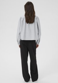 Person med langt brunt hår iført en lys grå langærmet bluse og løstsiddende sorte bukser, der står med ryggen til på en ensfarvet baggrund.