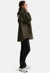 Femme aux cheveux tressés portant une veste à capuche vert olive, des leggings noirs et des baskets blanches, debout de profil sur fond blanc.