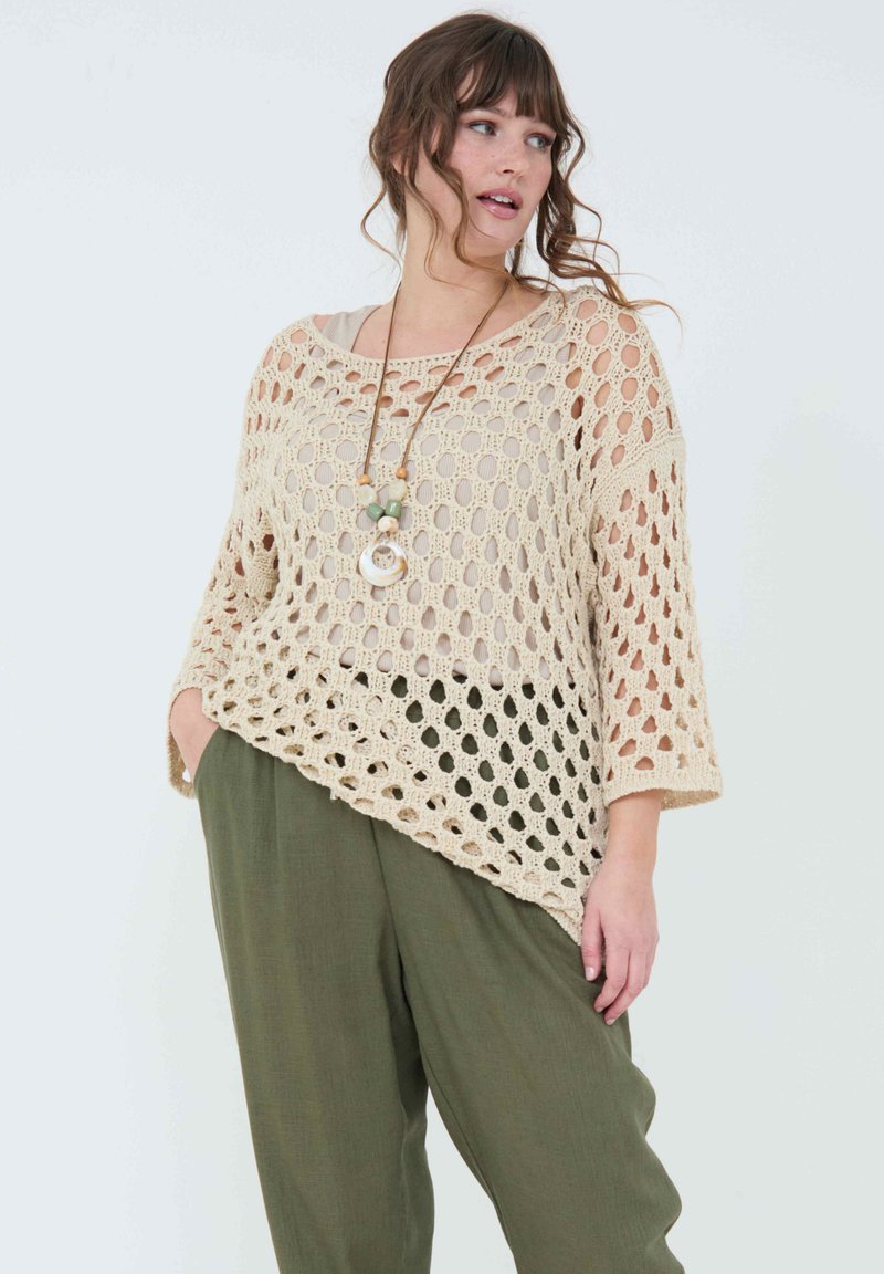 Un pull en maille ouverte beige clair avec un ourlet asymétrique, des manches trois-quarts et une coupe décontractée, associé à un pantalon ample en vert olive.