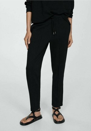 Person trägt schwarze, schmal zulaufende Hose mit Kordelzug in der Taille und schwarze Riemchensandalen, steht auf grauem Boden.
