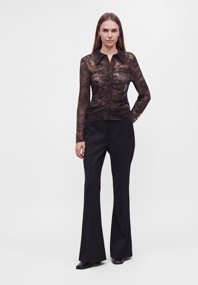 Femme portant un chemisier en dentelle noire transparente à boutonnières avec col, associé à un pantalon évasé noir taille haute et des chaussures noires.