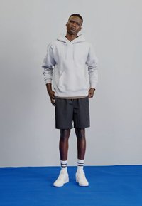 Jasnoszara bluza z kapturem z kieszenią kangurzą, czarne szorty oraz białe sneakerzy z paskowanymi skarpetkami; model stoi na niebieskiej powierzchni.