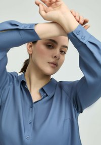 Young person with light skin and dark hair wearing a blue button-up shirt, raising crossed arms above head against a plain background.