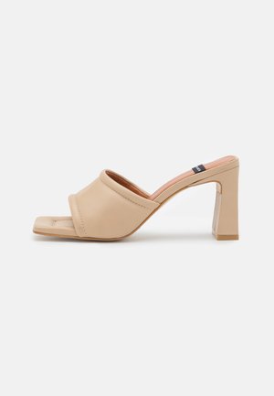 Beige open teen mule met gewatteerde brede band, vierkante blokhak en een klein zwart label aan de binnenzijde van de hak.