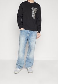 Sudadera negra con estampado gráfico en la parte frontal, combinada con jeans de pierna ancha de color azul claro y zapatillas grises. Estilo casual, diseño minimalista.