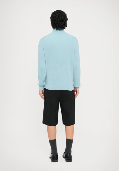 Pull côtelé bleu clair à col montant, associé à un short noir jusqu'aux genoux, des chaussettes grises et des chaussures noires. Design simple, sans motifs visibles.