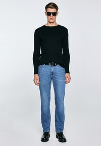 Mees, kes kannab musta pika varrukaga särki, siniseid teksaseid, musta vööd, musti kingi ja tumedaid päikeseprille, seisab ühevärvilise heleda tausta ees.