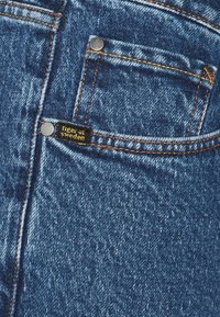 Close-up of blue denim jeans pocket with orange stitching and a small black label reading "tiger of sweden."