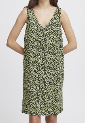 Robe sans manches à col en V avec fond noir et petit motif floral vert et blanc, portée par une femme les mains dans le dos.