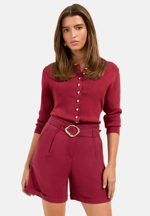 Žena oblečená v bordovom zapínanom kardigane so zlatými gombíkmi a k nemu sa hodia vysoké šortky s opaskom so zlatou sponou, stojaca pred bielym pozadím.