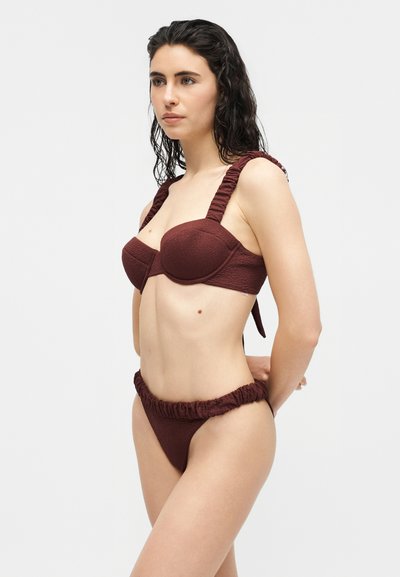 Femme aux cheveux noirs mouillés portant un bikini rouge foncé texturé avec des bretelles et une ceinture froncées, debout devant un fond clair uni.