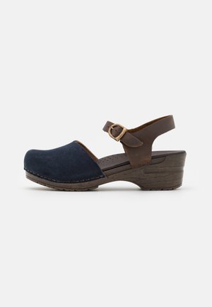 Sandale femme style sabot avec bout fermé en daim bleu marine, bride de cheville en cuir marron avec boucle en laiton, et petit talon bloc en bois.