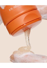 Klar geléaktig produkt i en oransje beholder, dosert på en hånd med skummende tekstur, mot en nøytral bakgrunn.