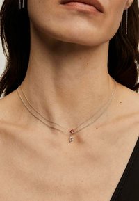 Collar de plata con capas que presenta un acento rojo y un pequeño charm con la letra "P", exhibido en un cuello desnudo con mínimos detalles alrededor.